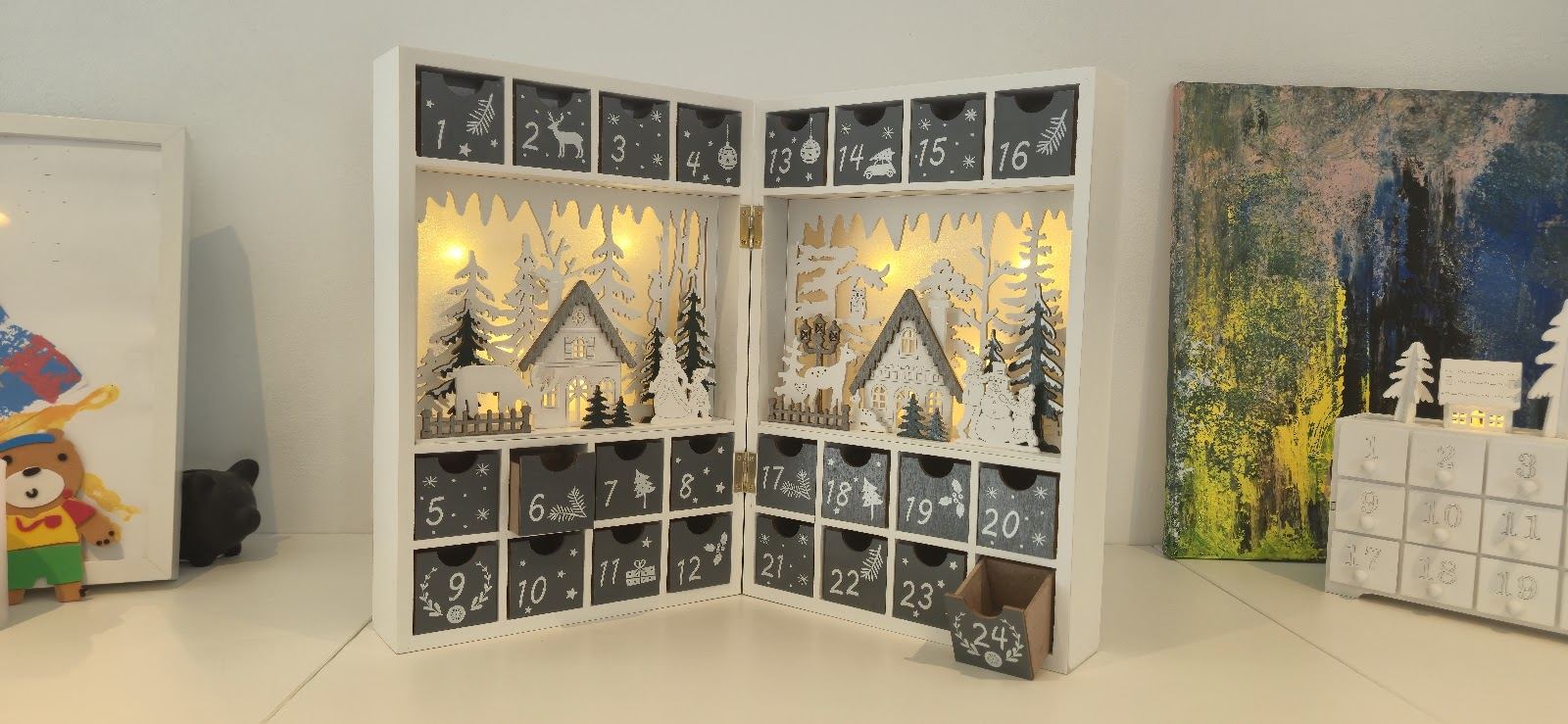 Osvětlený dřevěný adventní kalendář s malými šuplíčky a vyřezávanou zimní krajinou.